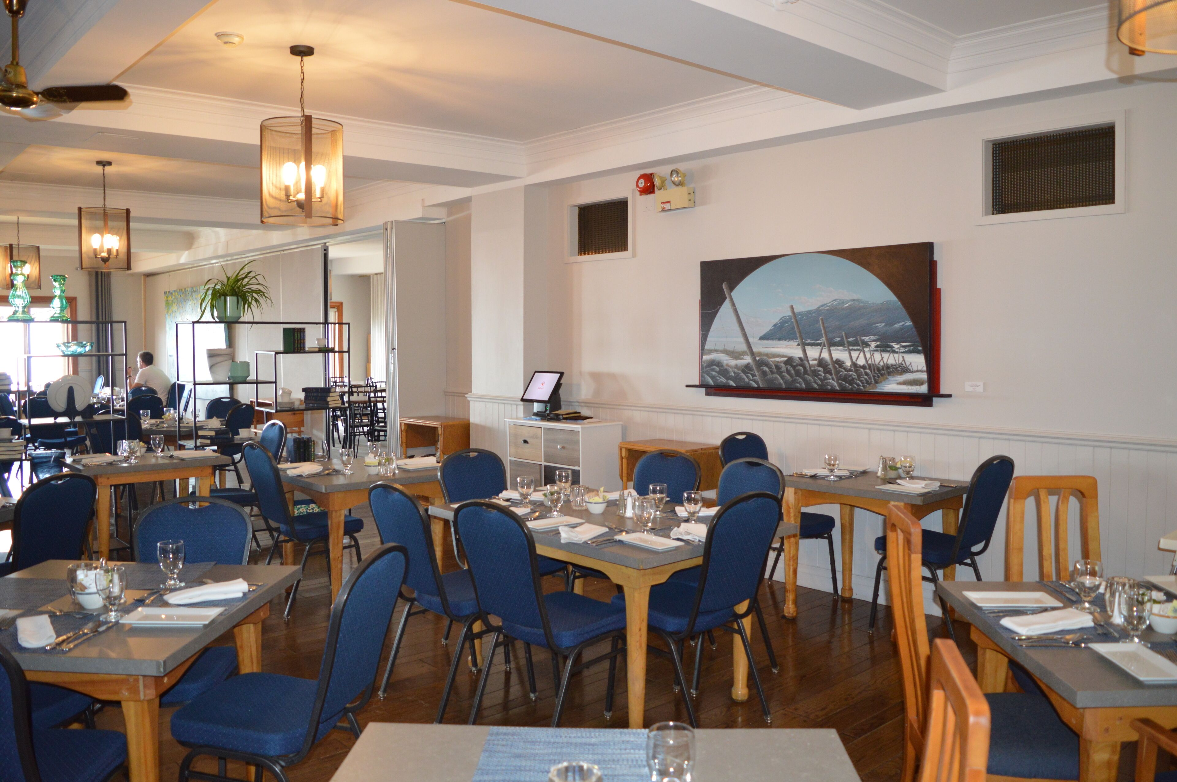 A restaurant with multiple tables and chairs, a ceiling fan, and a painting on the wall.