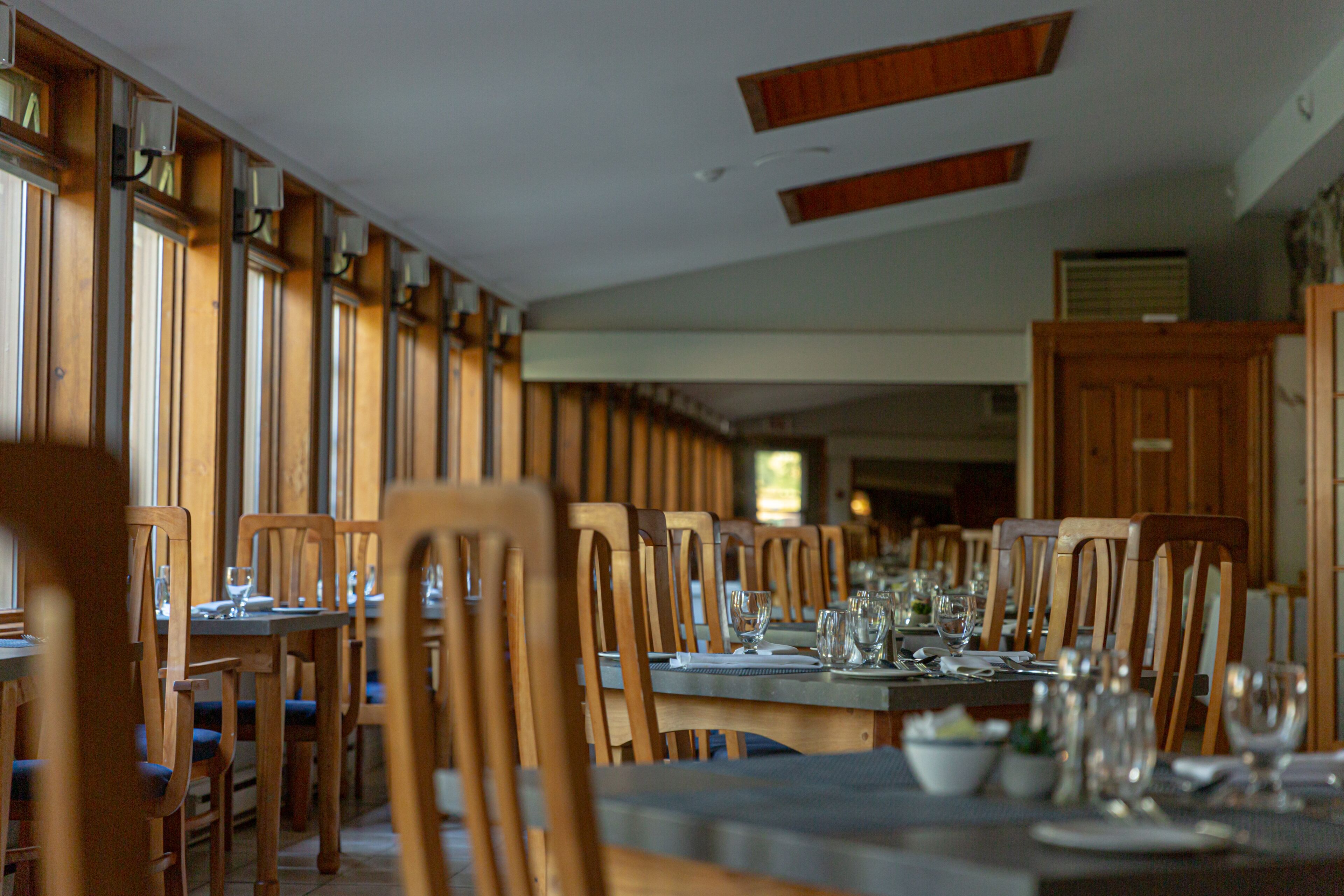 Un restaurante con sillas y mesas de madera, ventanas amplias y un techo alto con vigas de madera.