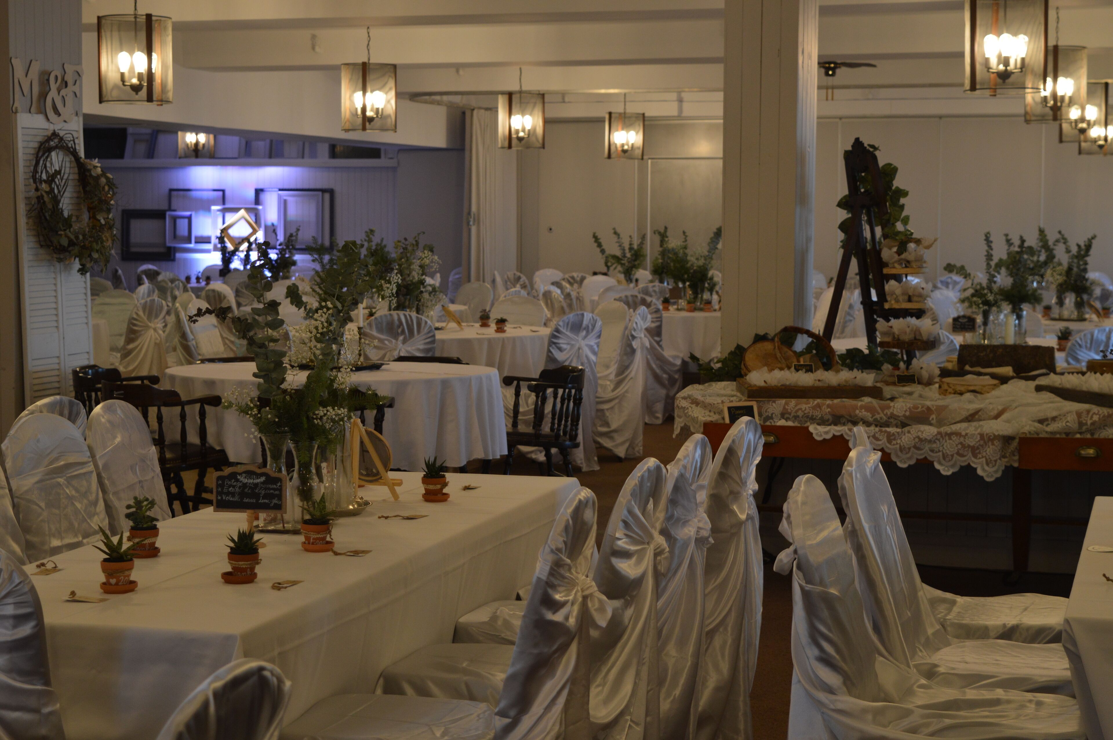 Un salón de banquetes con mesas dispuestas para un evento formal, cubiertas con manteles blancos y decoradas con plantas en macetas.