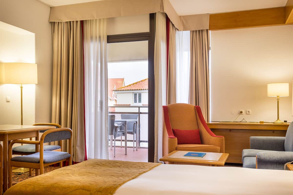 Habitación de hotel con cama, dos sillones, una mesita y vistas al balcón exterior.