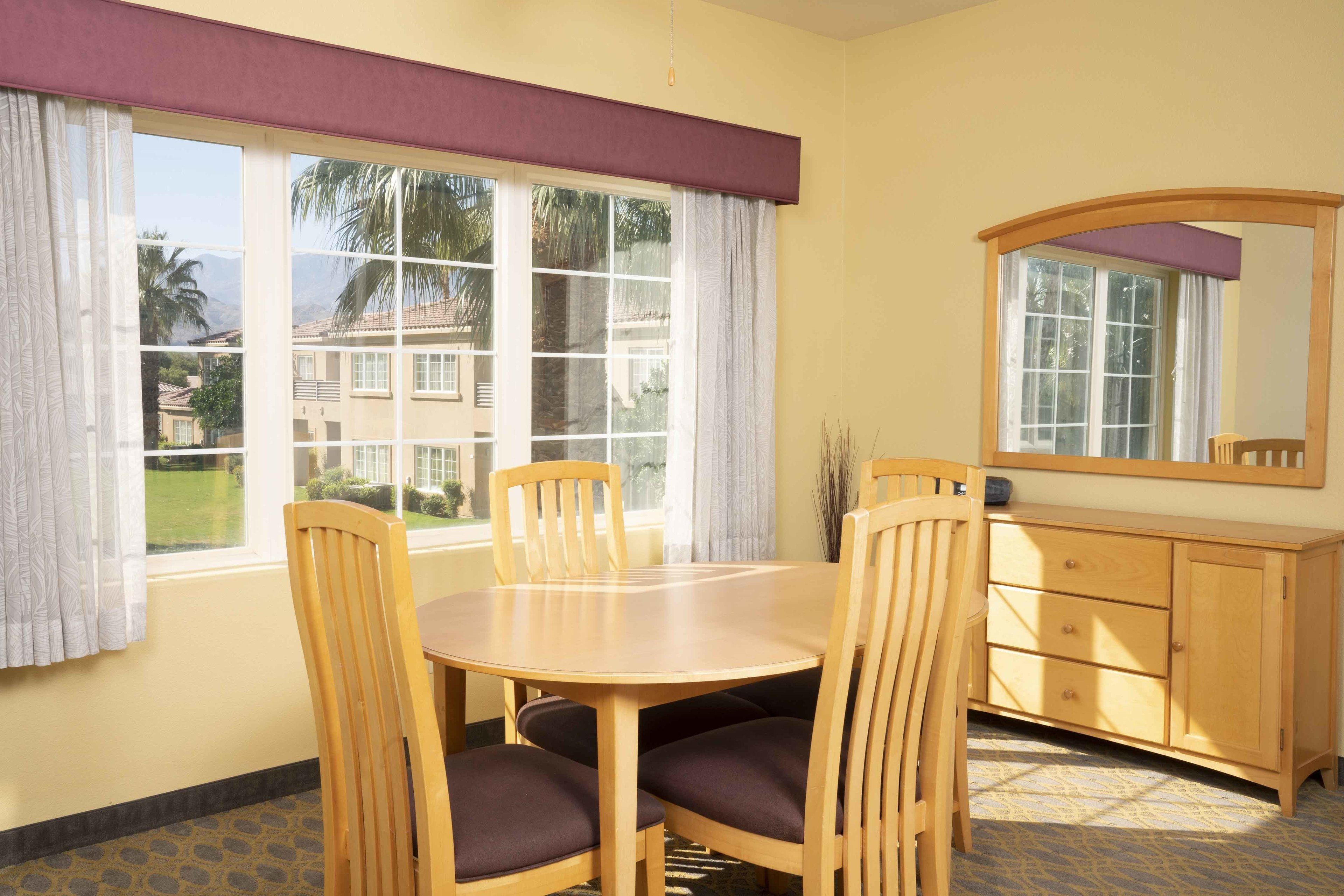 A room with a round wooden table and four chairs, a wooden cabinet with drawers, and a large window with curtains.