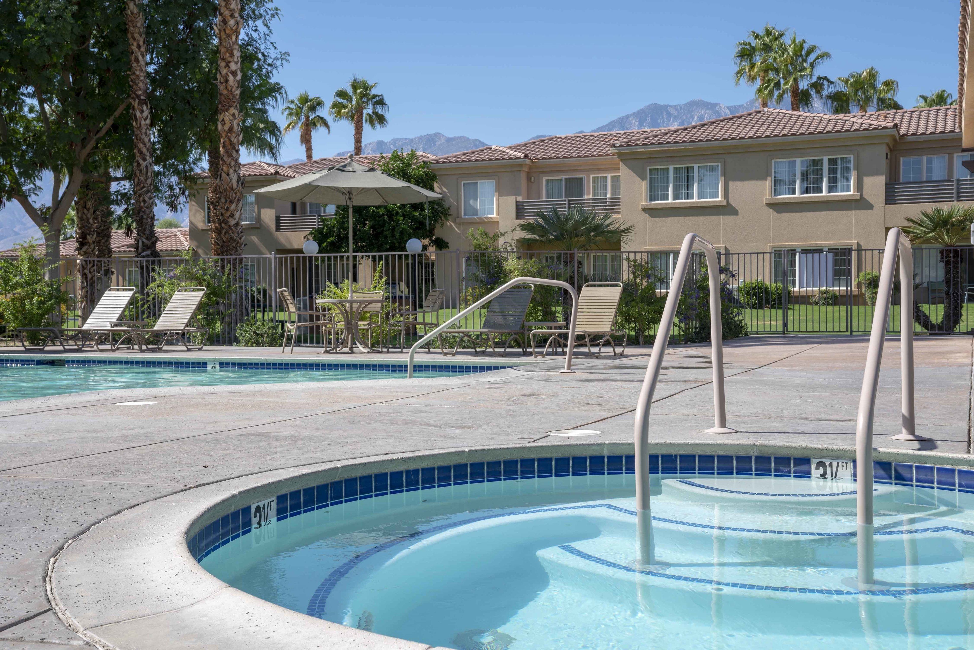 A swimming pool area with lounge chairs, a hot tub, and a building with a tiled roof in the background.
