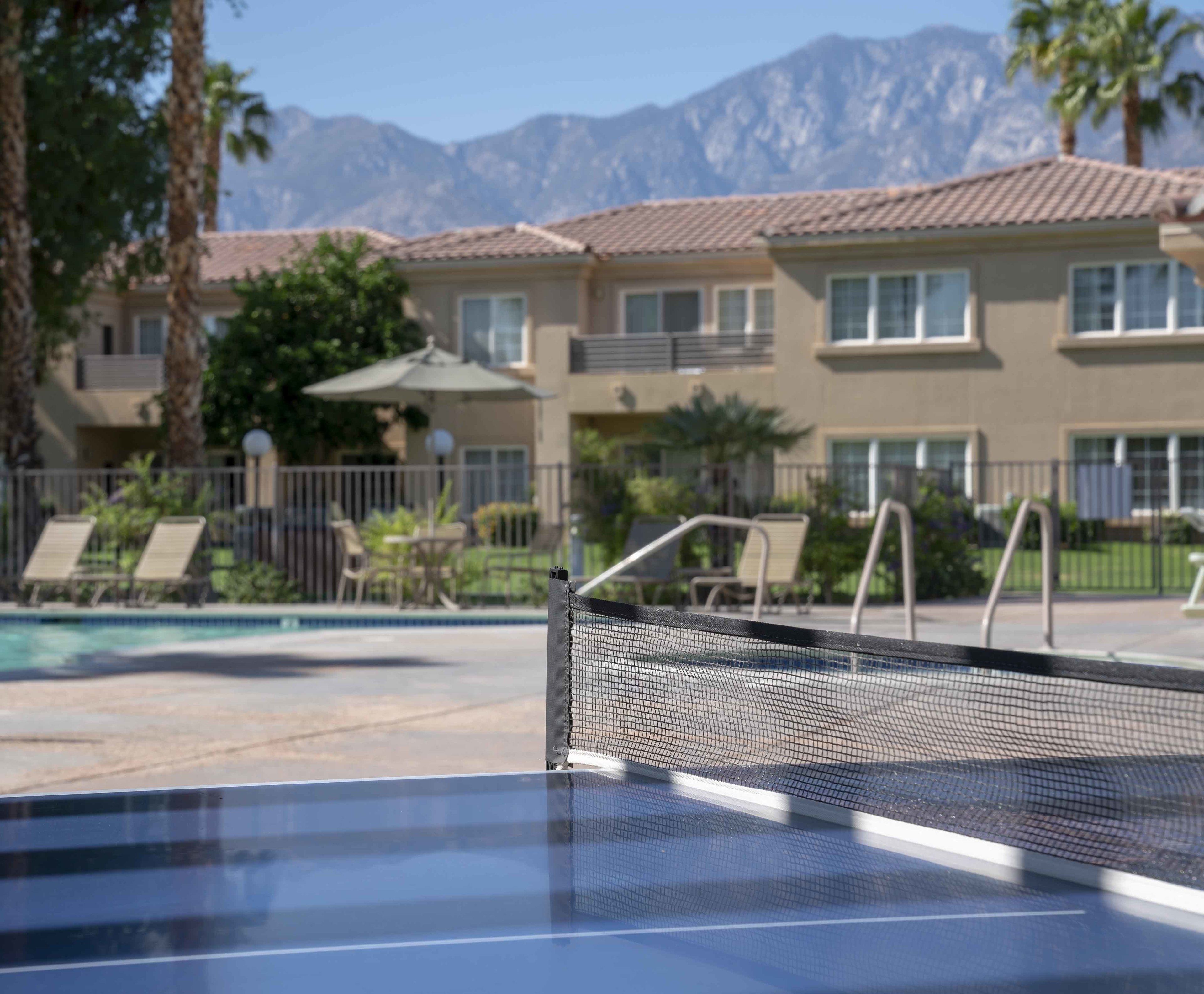 A pool area with a netted tennis court, lounge chairs, and a view of multi-story buildings with a mountainous backdrop.