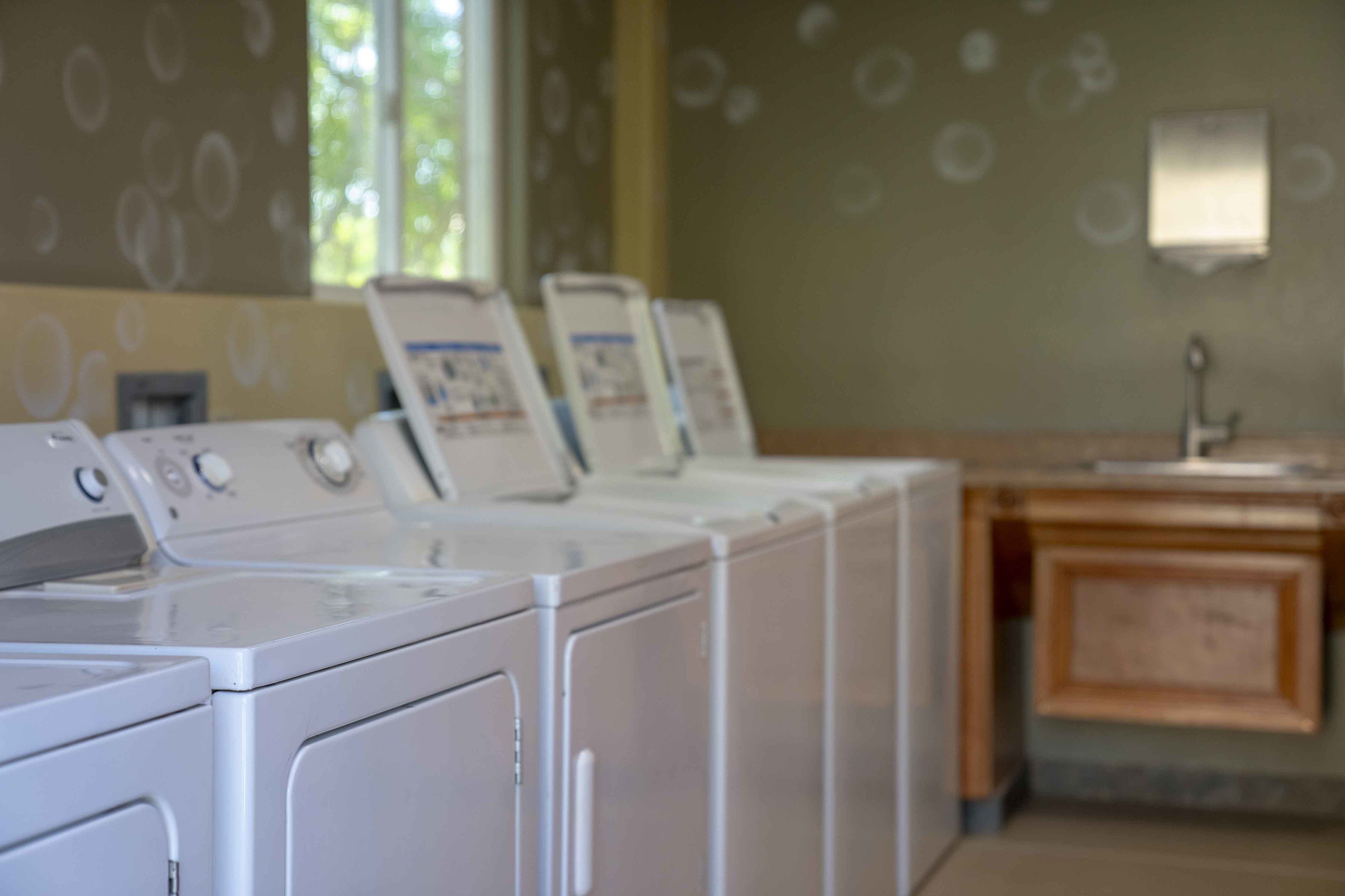 A row of white front-loading washers in a laundry room with a wooden cabinet and a wall-mounted light fixture.