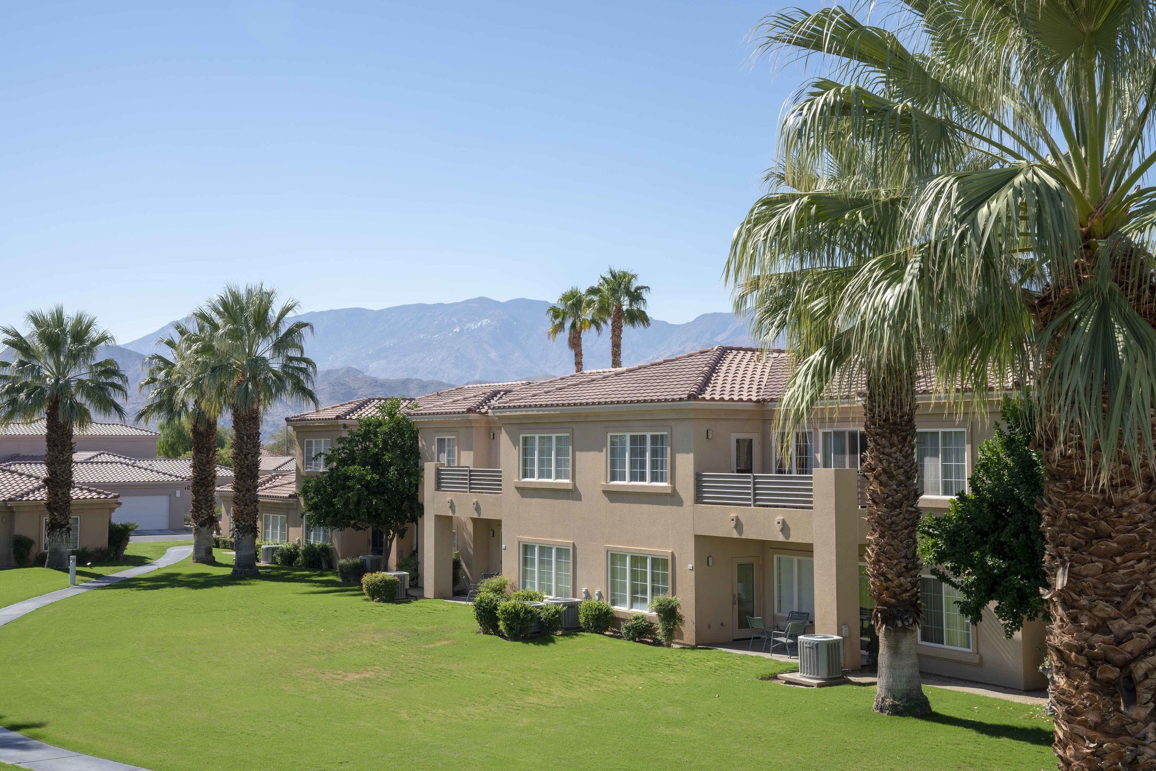 A multi-story residential building with a flat roof, surrounded by well-maintained lawns and palm trees.