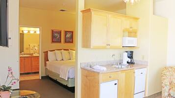 Habitación de hotel con cama, una pequeña zona de estar con mesa y sillón, kitchenette con microondas y cafetera, y baño con lavamanos y espejo.