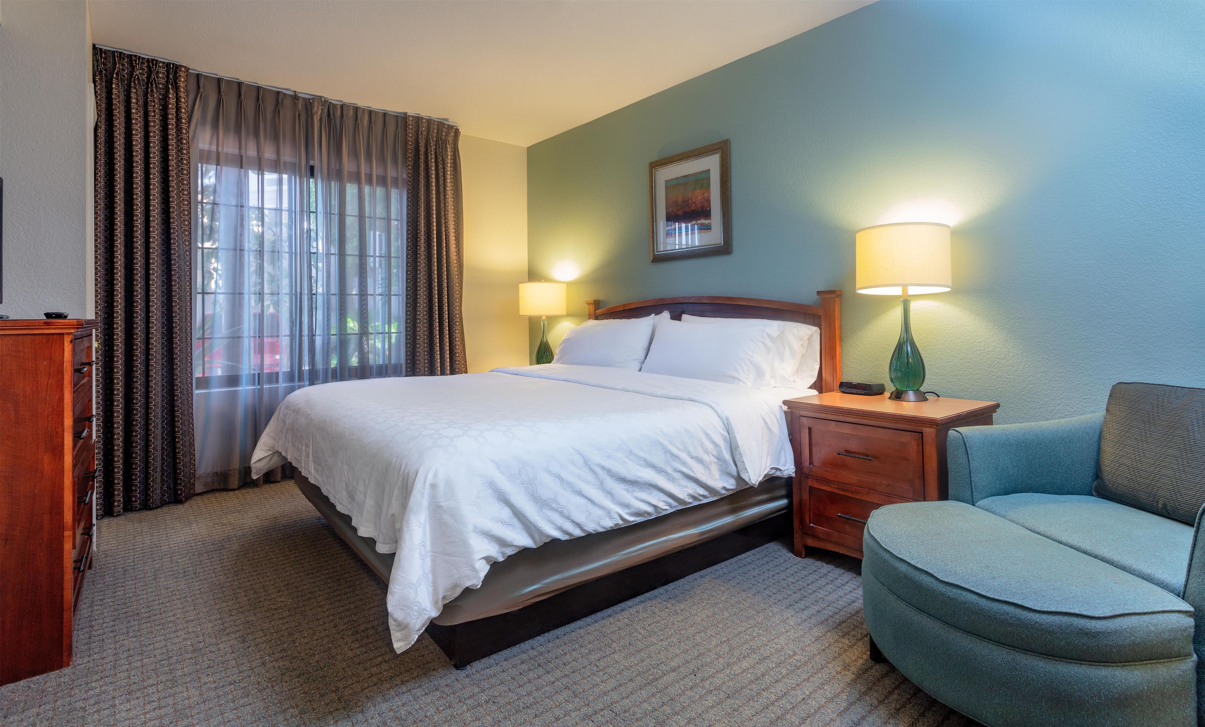 Quarto de hotel com cama, duas mesas de cabeceira com abajures, uma cadeira, cômoda e janela com cortinas.