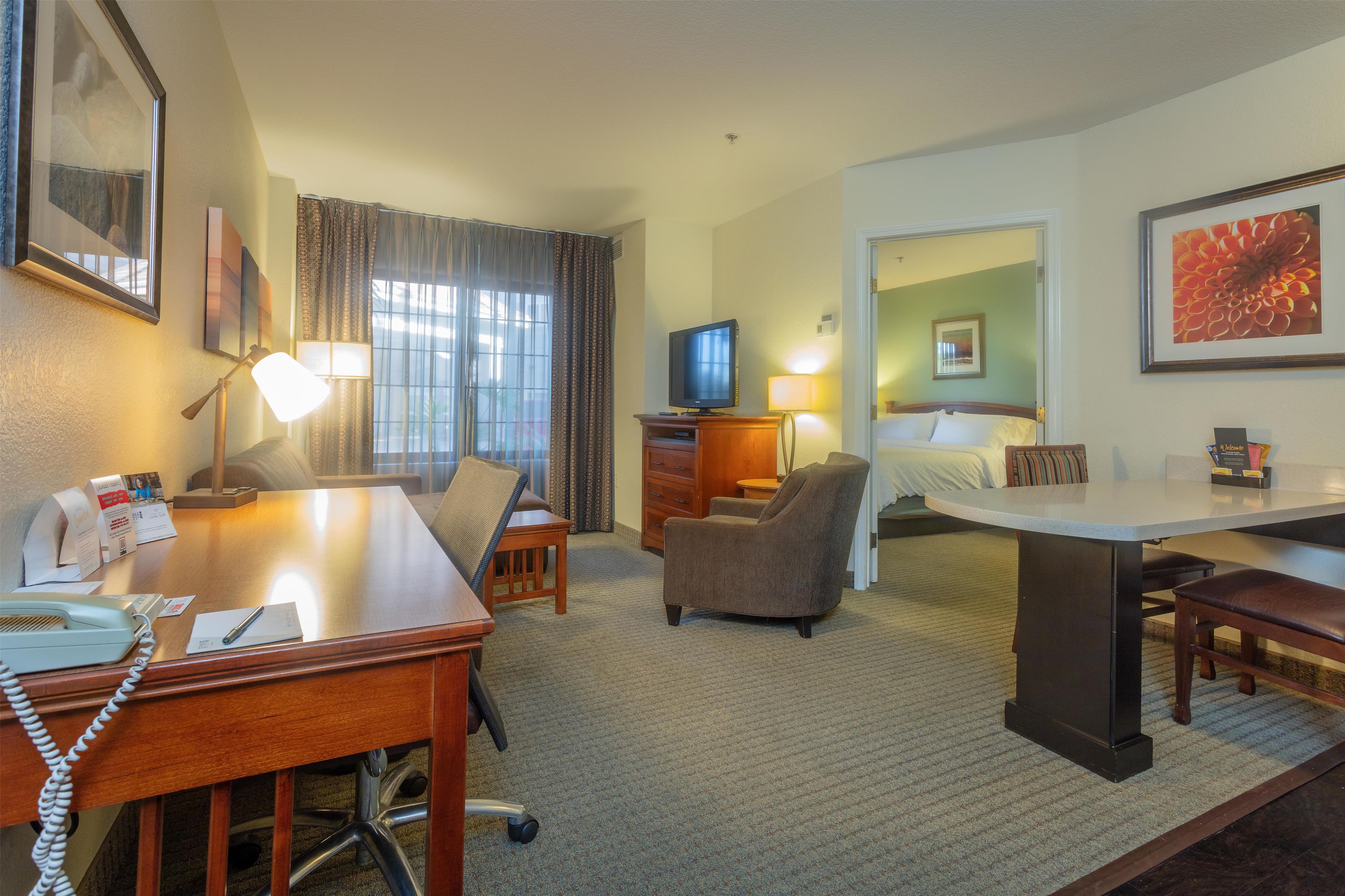 A hotel room with a desk, chair, television, bed, and a large window with curtains.