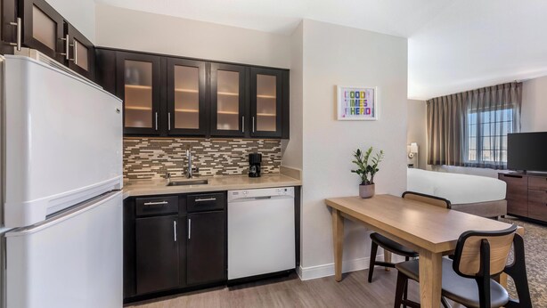Una cocina compacta con armarios oscuros, un refrigerador blanco y un lavavajillas. Una zona de comedor con una mesa y sillas de madera está contigua a la cocina.