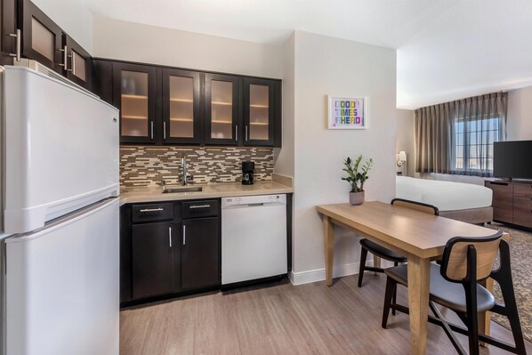 A compact kitchen with dark cabinets, a white refrigerator, and a dishwasher. A dining area with a wooden table and chairs is adjacent to the kitchen. - Sonesta ES Suites Anaheim Resort Area (Anaheim)