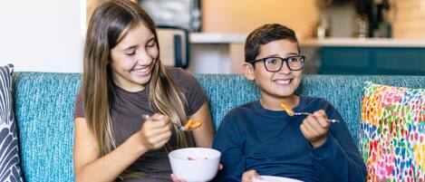 Two people sitting on a teal sofa with a colorful pillow, eating from white bowls.
