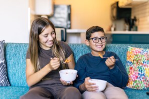 Two people sitting on a teal sofa with a colorful pillow, eating from white bowls. - Sonesta ES Suites Anaheim Resort Area (Anaheim)