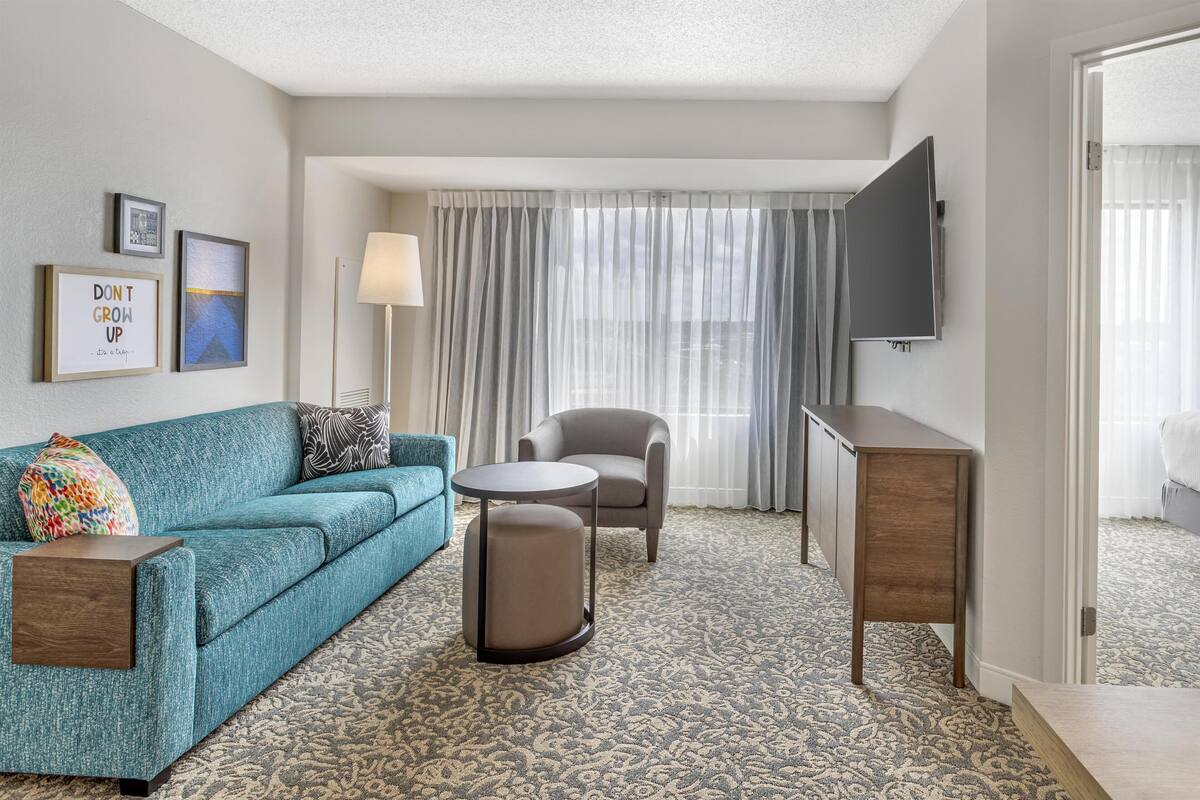 Ein Hotelzimmer mit einem türkisfarbenen Sofa, einem runden Hocker, einem grauen Sessel, einem kleinen Tisch, einem an der Wand montierten Fernseher und einem gemusterten Teppich.