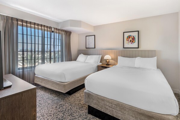 Habitación de hotel con dos camas, un escritorio, una lámpara y un ventanal grande con cortinas.