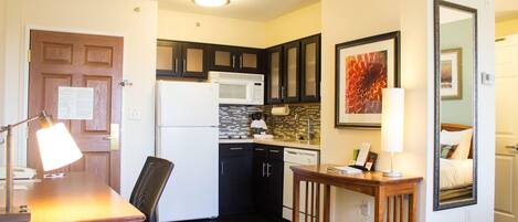 A compact kitchen with a refrigerator, microwave, and dishwasher. There is a dining table with a chair and a lamp. A mirror is on the wall, and a framed picture is above it.