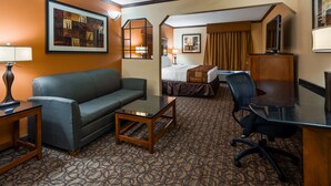 A hotel room with a sofa, a small table, a desk with a chair, a bed with a headboard, a television, and a painting on the wall. - Best Western Plus Midwest Inn & Suites (Salina)