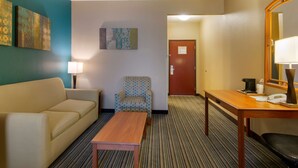 A hotel room with a sofa, armchair, small table, lamp, and a door with a sign.