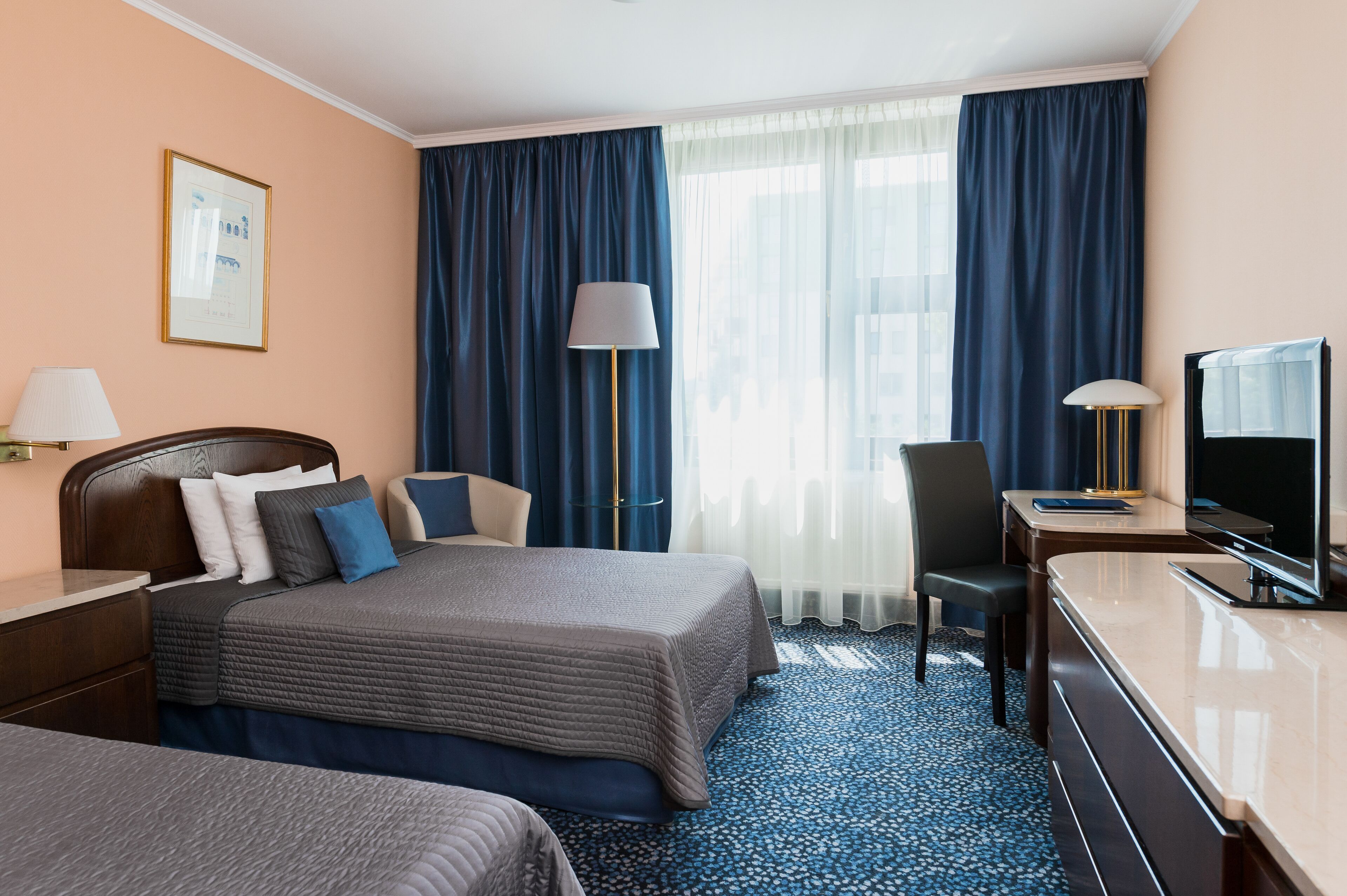 A hotel room with a bed, two bedside tables, a desk with a TV, a chair, and a large window with curtains.