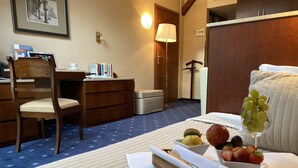 Ett hotellrum med en säng, ett skrivbord, en stol och en matplats med frukt och ett vinglas.