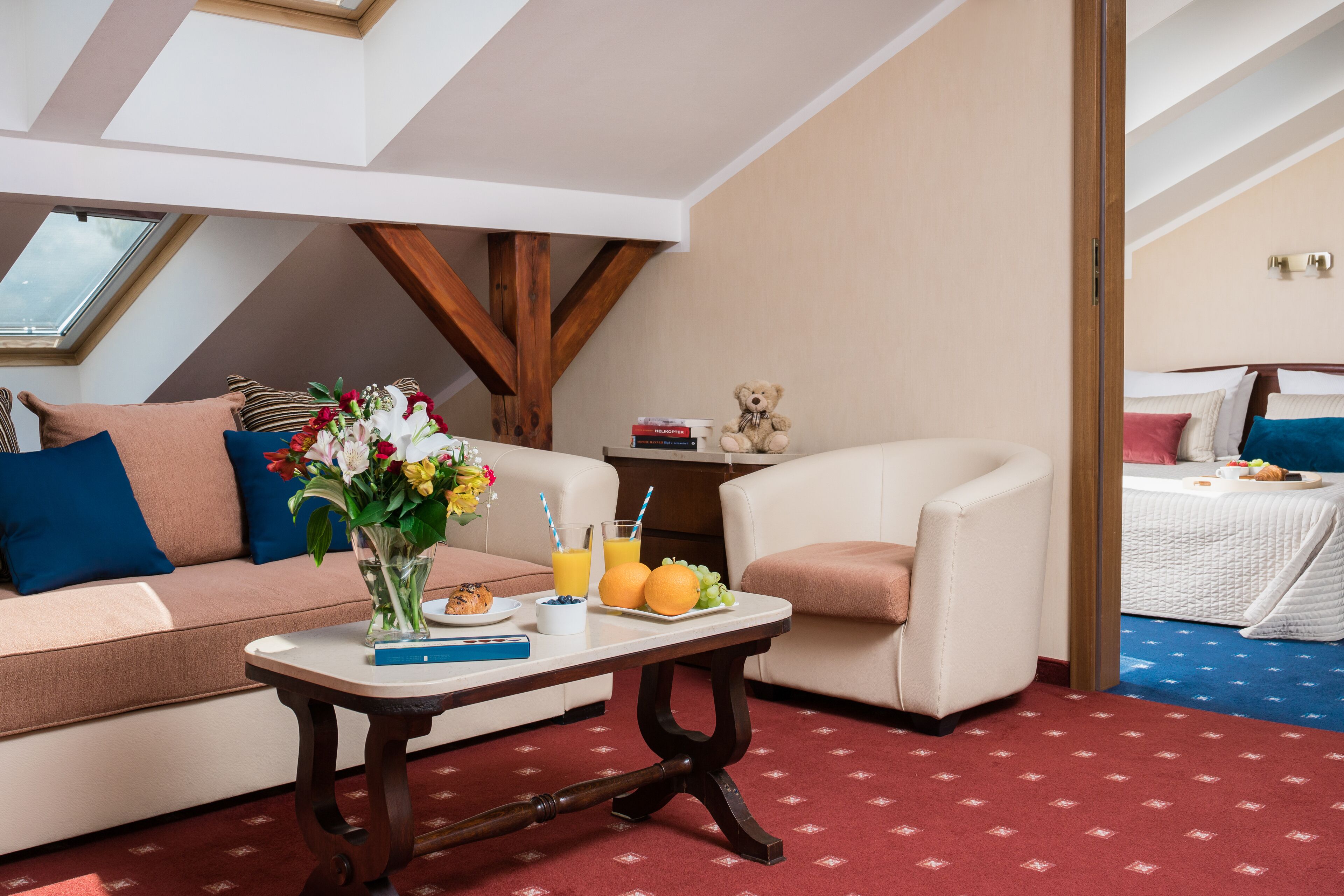 Una habitación acogedora con sofá, una mesita con un jarrón de flores, un osito de peluche y una cama con edredón azul y blanco.