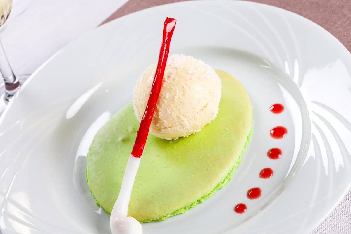 A dessert on a white plate with a green base, a white sphere on top, and a red and white striped item.