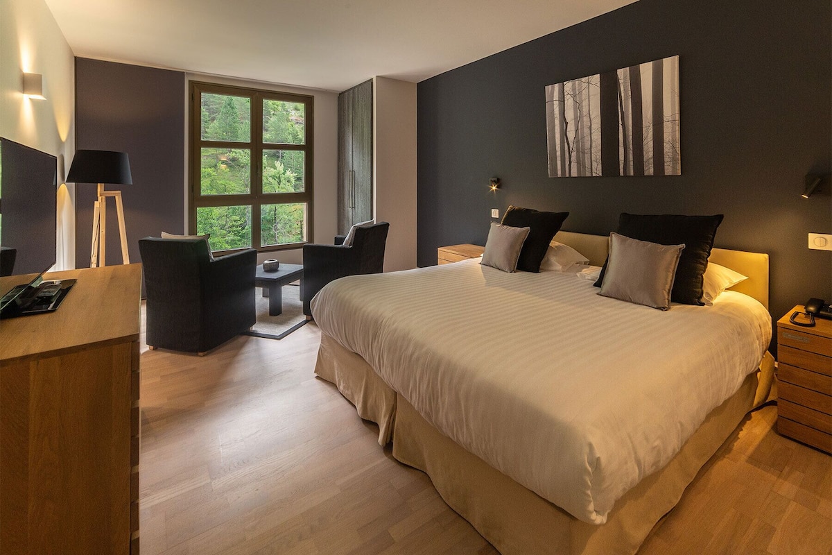 A modern bedroom with a large bed, a desk, and a chair. There is a window with a view of trees.