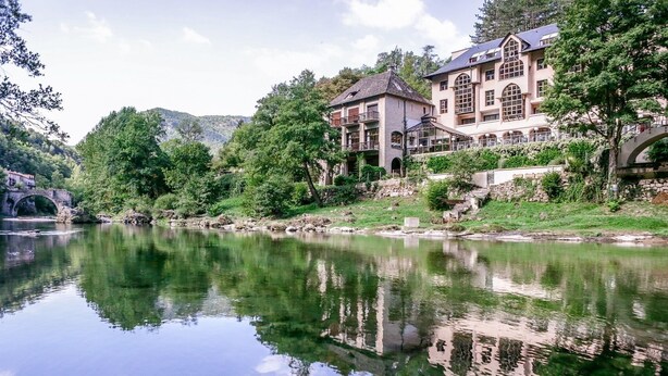 Un edificio grande e histórico de varios pisos y con un balcón, ubicado junto a un río tranquilo con un puente de piedra al fondo.