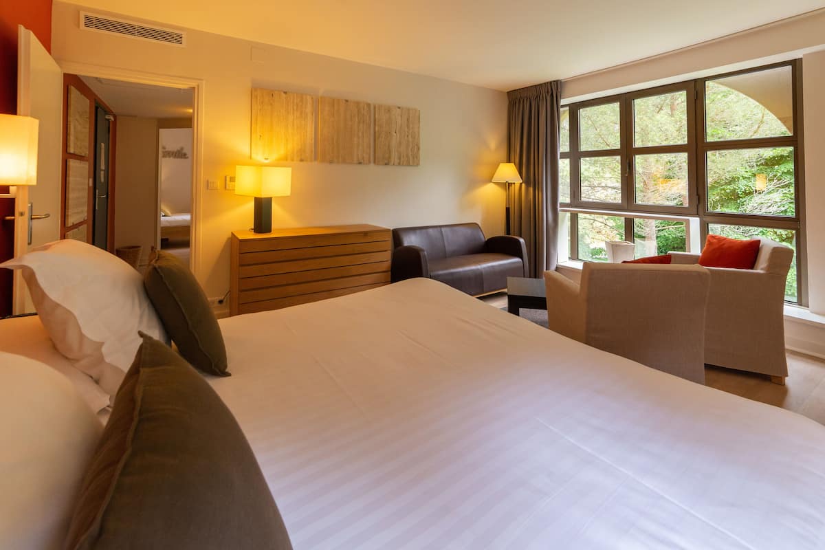 A hotel room with a large bed, a desk, a chair, and a window with a view of greenery.