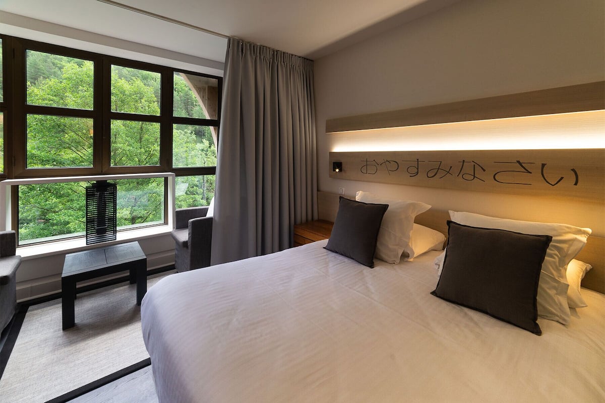 A modern hotel room with a large bed, a desk, and a view of greenery through the windows.