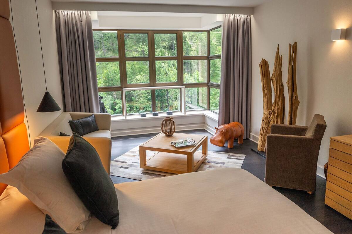 A modern hotel room with a large window, a sofa, a bed, a coffee table, and a decorative wooden sculpture.