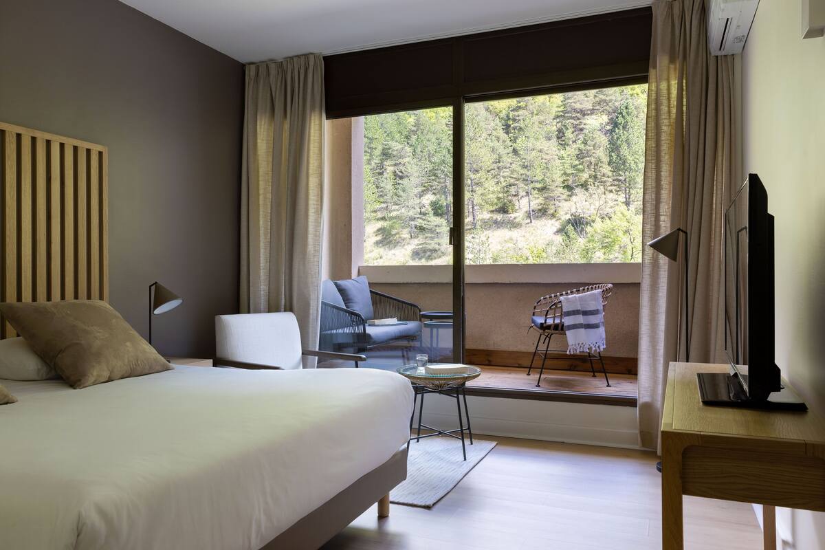 A bedroom with a bed, a chair, a small table, a TV, and a window with a view of trees.