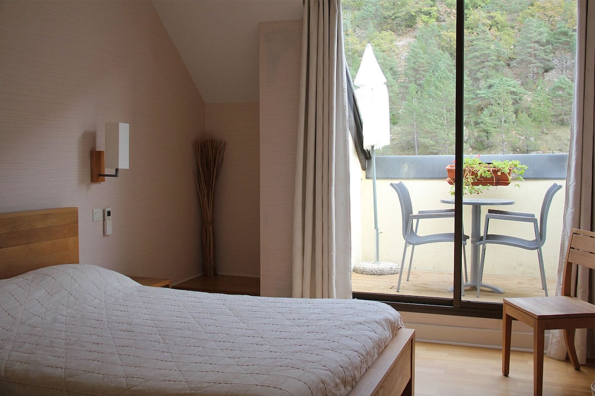 A bedroom with a bed, a bedside table, a window with curtains, and a view of an outdoor patio with chairs and a table.