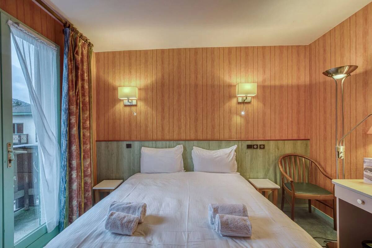A hotel room with a large bed, two wall-mounted lamps, a wooden headboard, a wooden desk with a chair, and a view through a glass door.