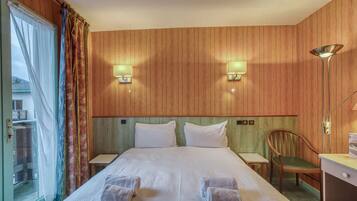 A hotel room with a large bed, two wall-mounted lamps, a wooden headboard, a wooden desk with a chair, and a view through a glass door.