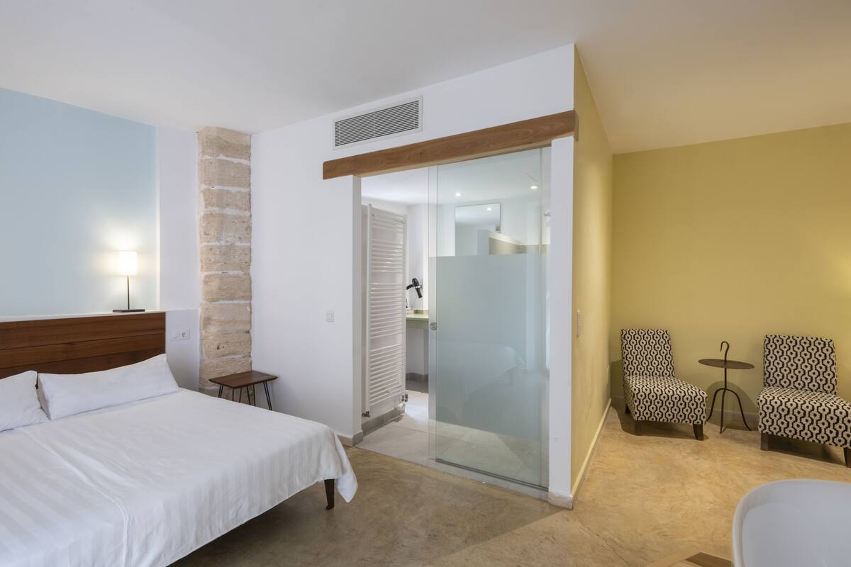 Une chambre d’hôtel avec un lit, une petite table, deux chaises à motifs, une porte en verre donnant sur une salle de bain et une lampe.