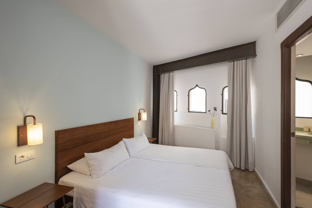 Une chambre d’hôtel avec un grand lit, une table de chevet, une lampe fixée au mur et une fenêtre avec des rideaux.