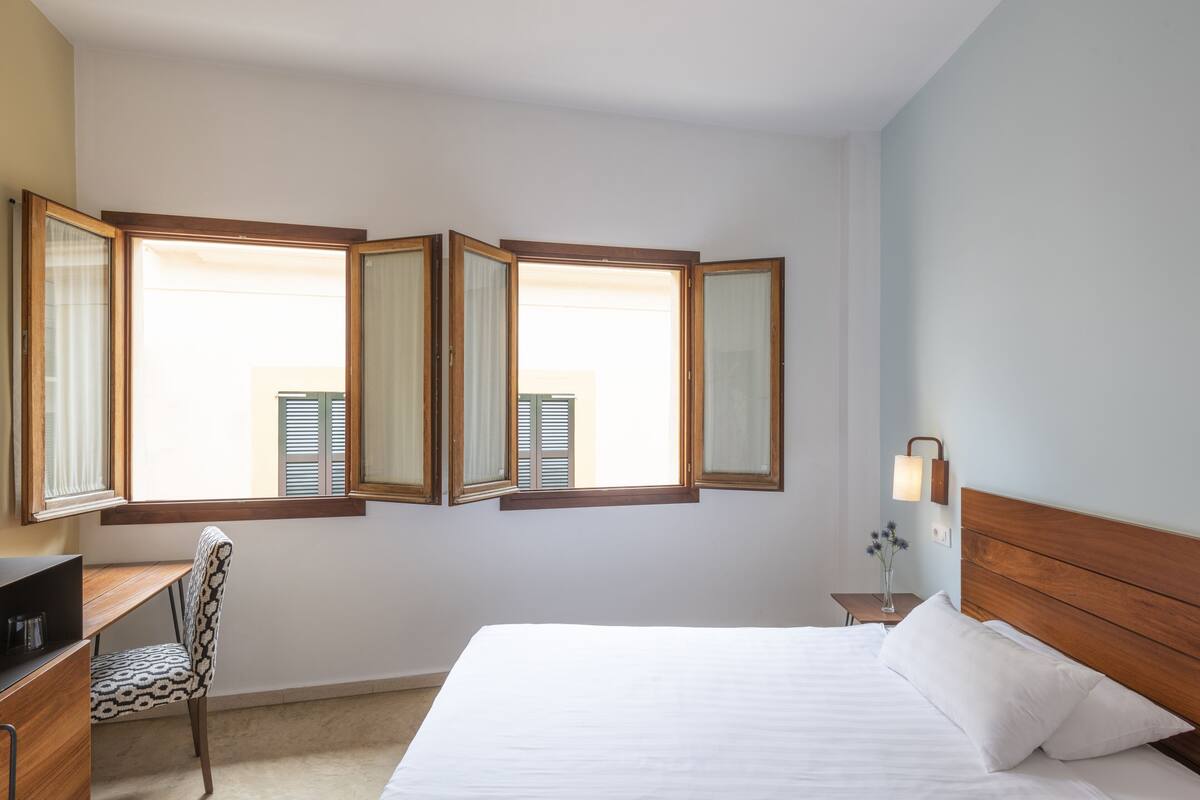 Une chambre à coucher avec un lit, un bureau et une chaise. Il y a trois fenêtres avec des rideaux.