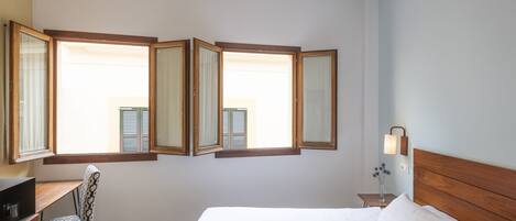 A bedroom with a bed, a desk, and a chair. There are three windows with curtains.