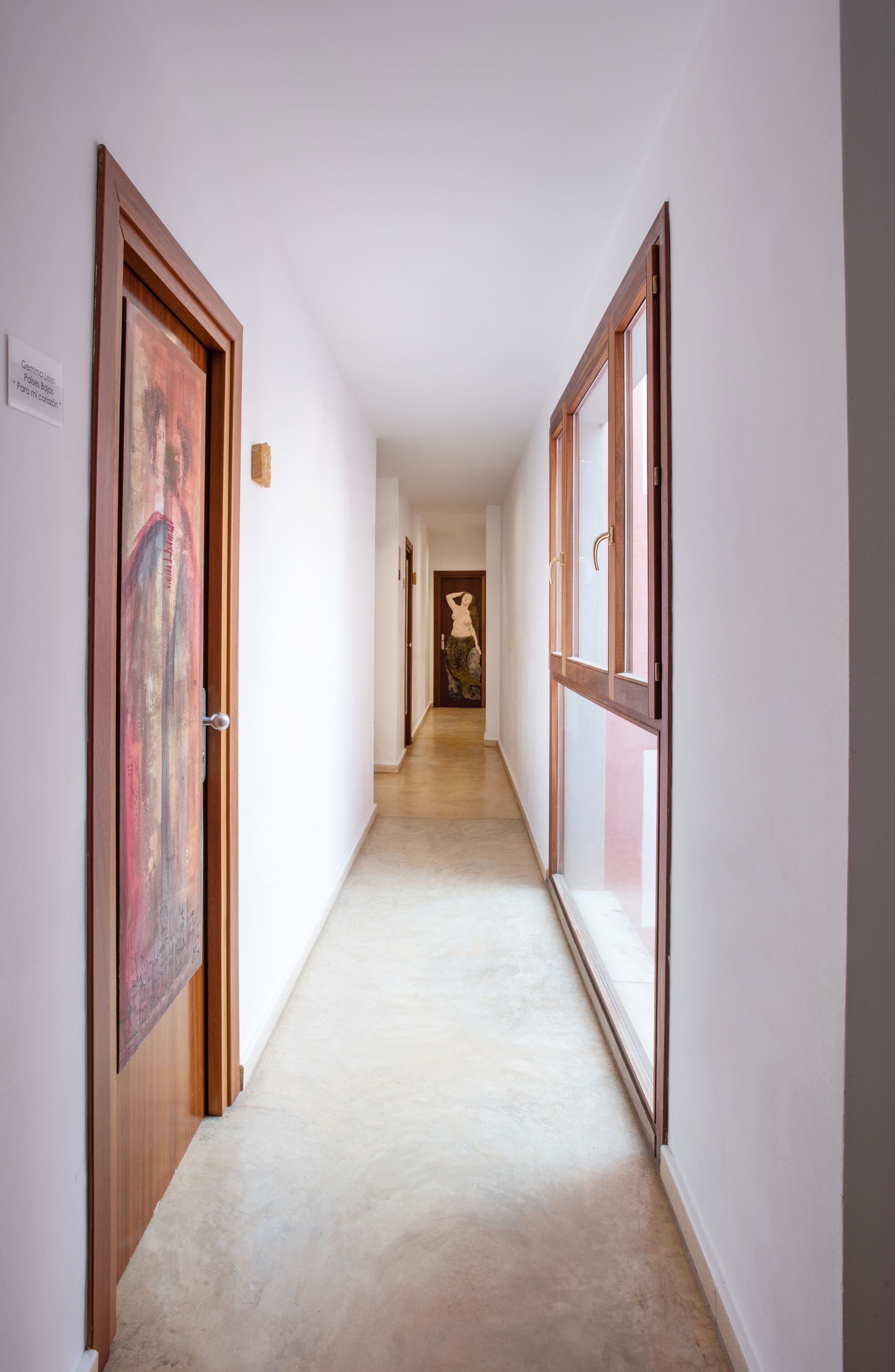A hallway with wooden doors and windows, leading to a room with a bed and a nightstand.
