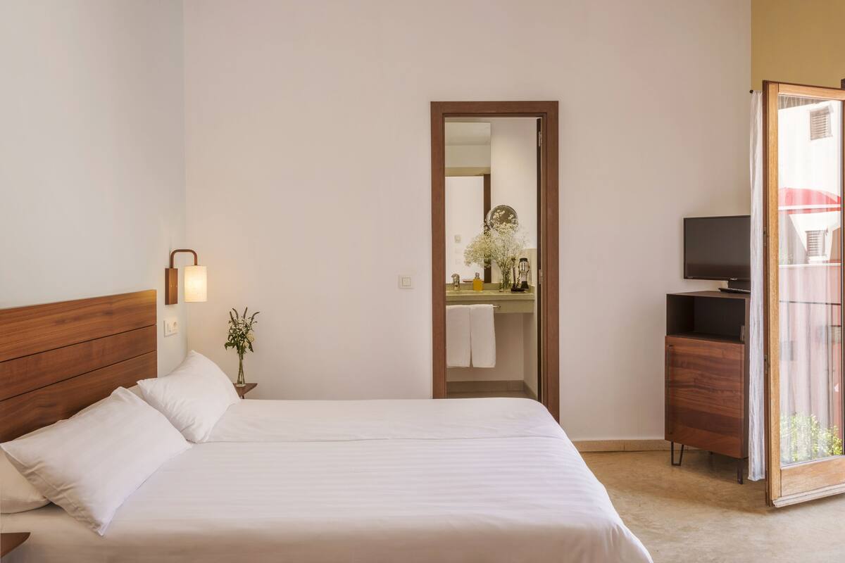 Une chambre d’hôtel avec un lit doté d’une tête de lit en bois, une lampe de chevet, une petite plante en pot, une salle de bain avec un miroir, un lavabo et un téléviseur posé sur un meuble en bois.
