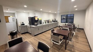 A modern kitchen with a coffee machine, a refrigerator, and a window with a view. There are tables and chairs arranged for dining.