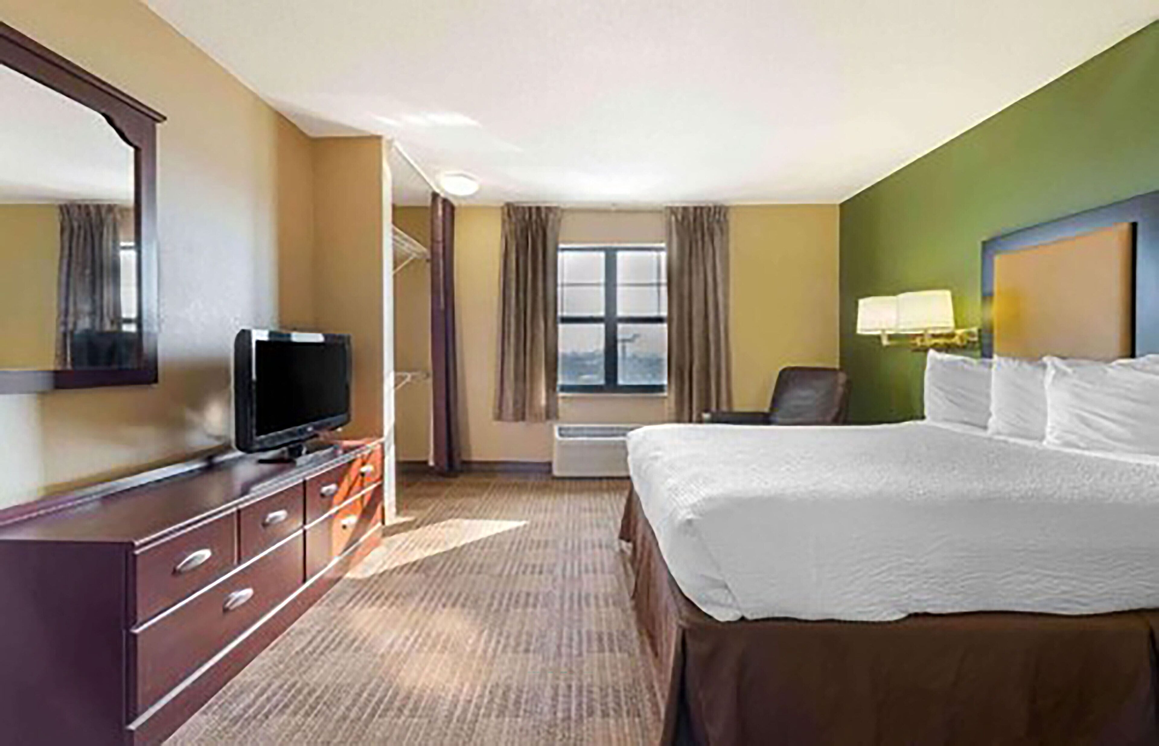 A hotel room with a large bed, a television on a dresser, a chair, a window with curtains, and a wall-mounted lamp.