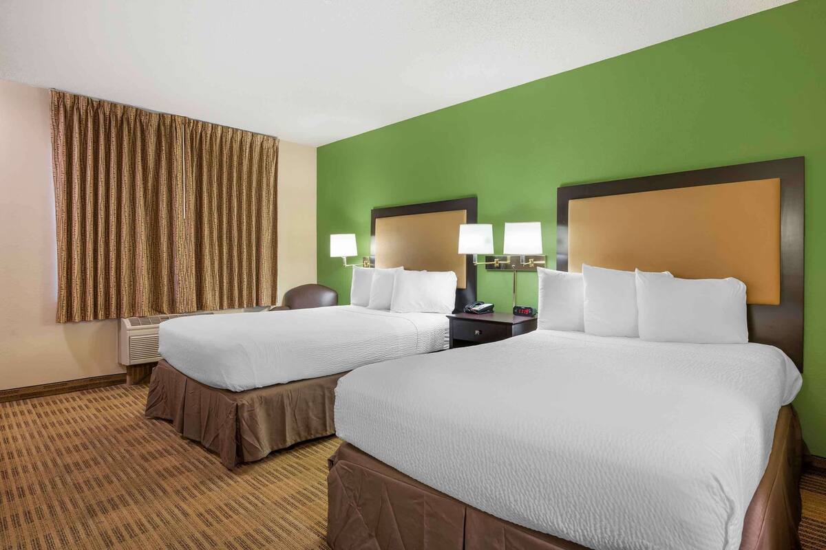 Habitación de hotel con dos camas, una pared de acento verde, un ventanal con cortinas y una mesita de noche con lámparas.