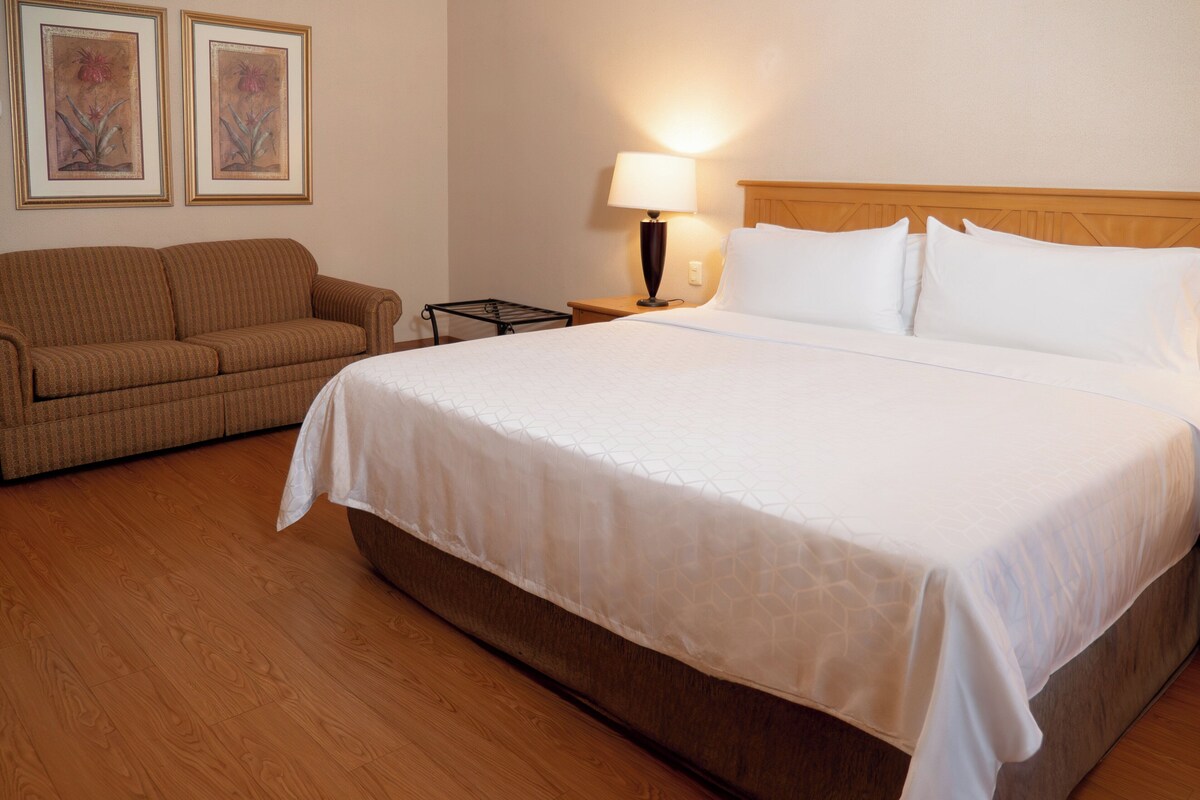 A hotel room with a large bed, a wooden headboard, a sofa, a nightstand with a lamp, and two framed pictures on the wall.