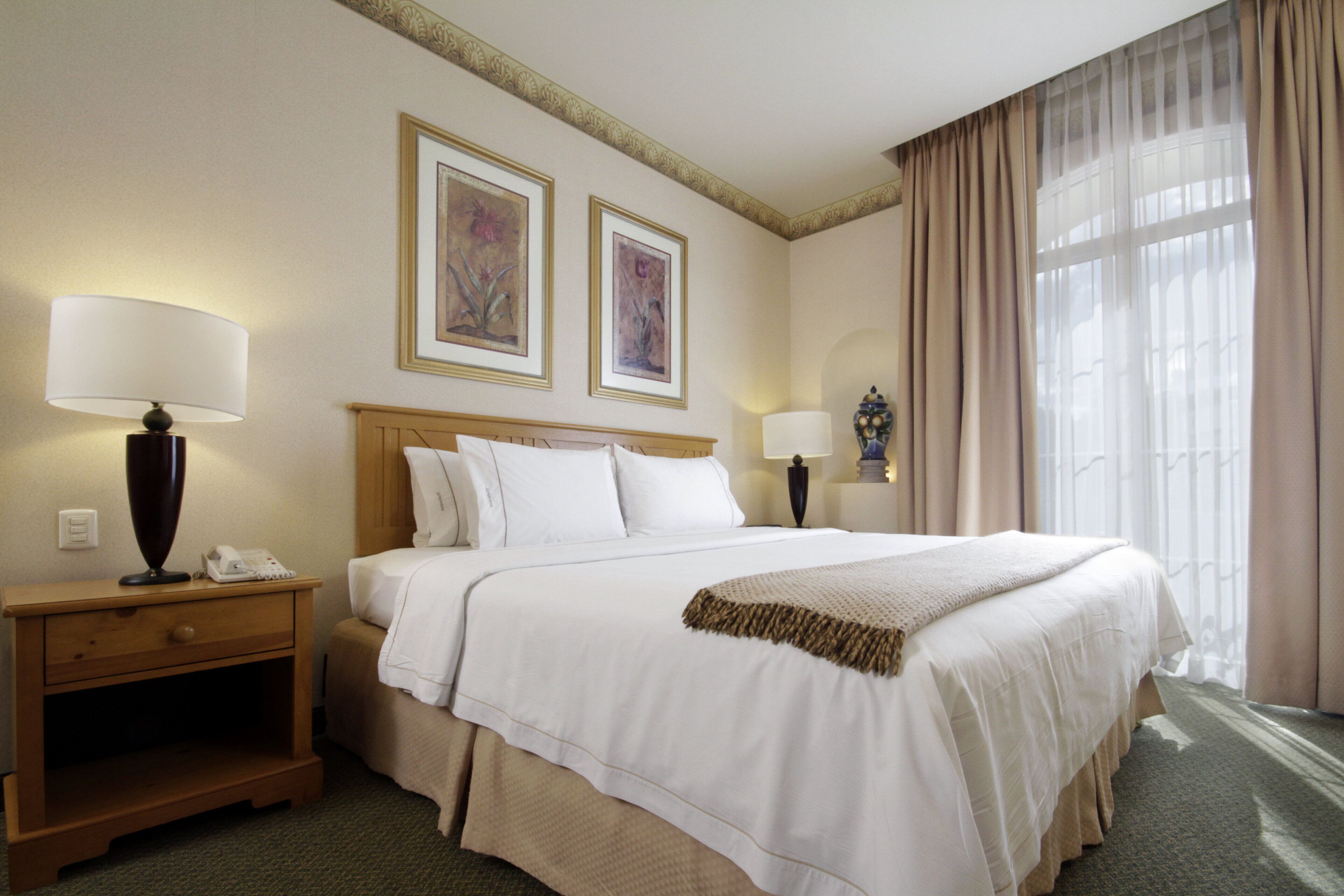 Habitación de hotel con una cama grande, dos lámparas de noche, una mesita de noche, un teléfono y dos cuadros en la pared.