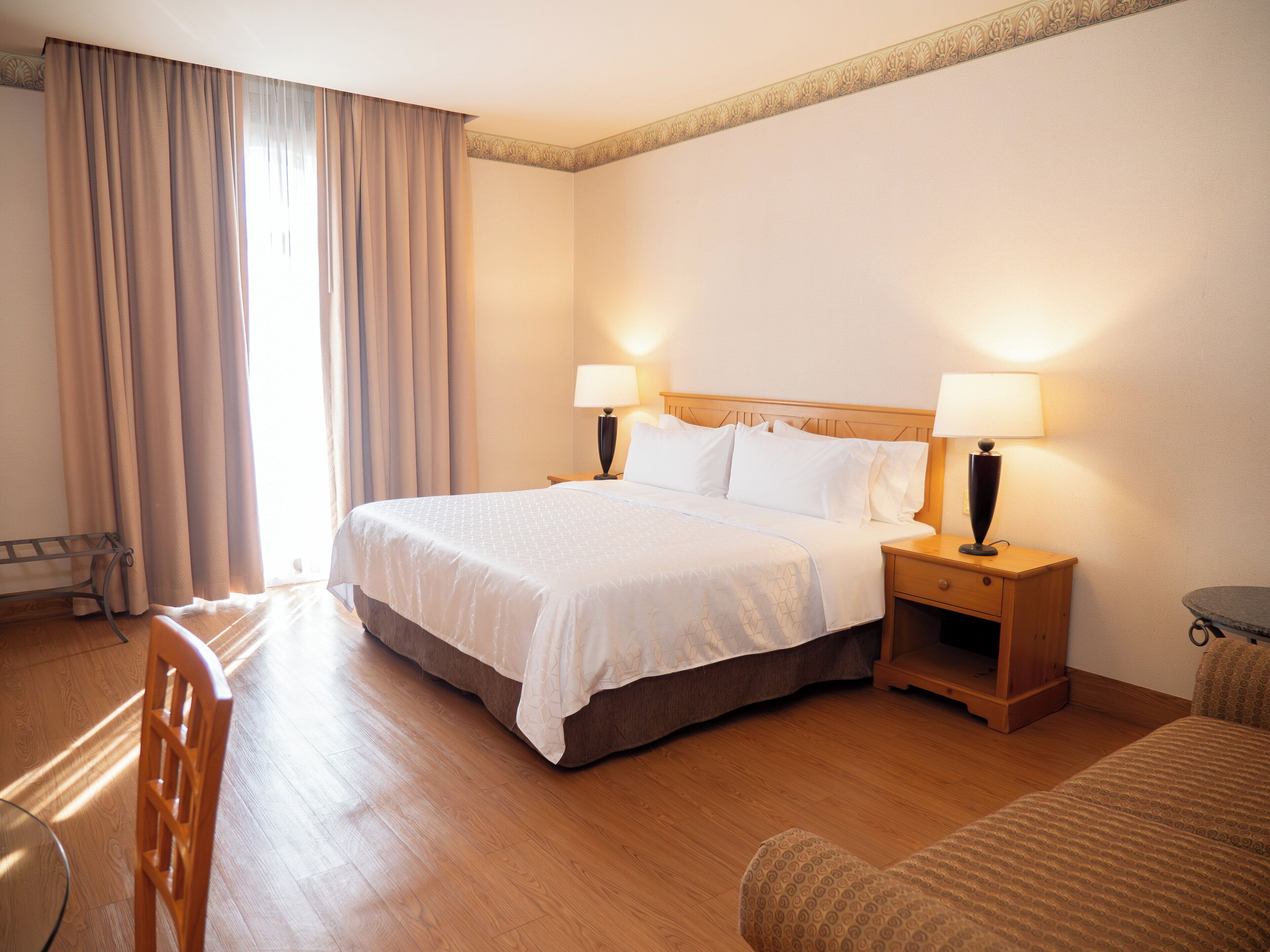 Una habitación de hotel con una cama grande, dos mesitas de noche con lámparas, una silla de madera y una pequeña mesa redonda.