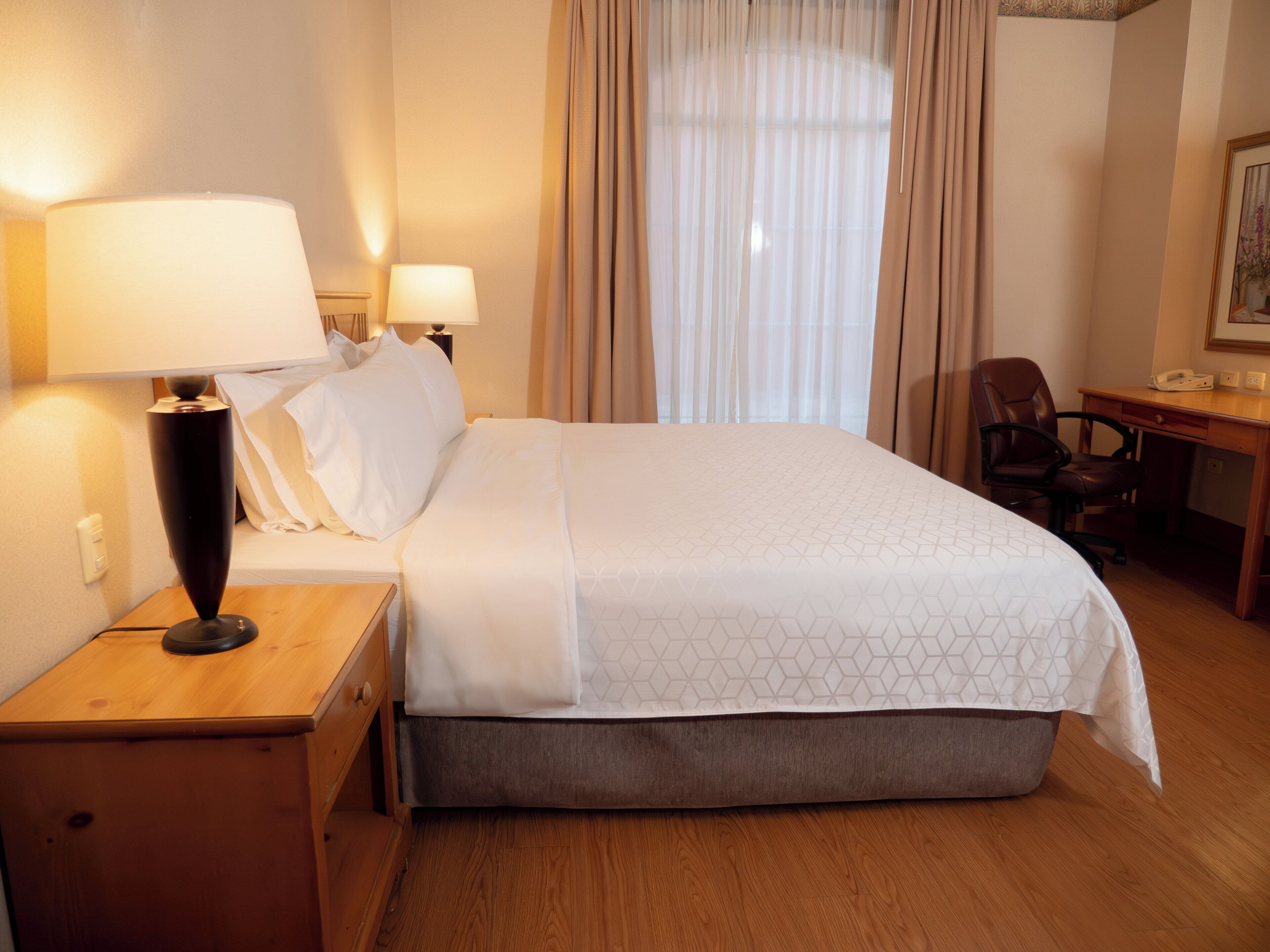 A hotel room with a bed, two bedside lamps, a desk with a chair, and a window with curtains.