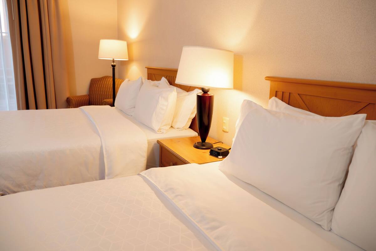 Una habitación de hotel con dos camas, una mesita de noche con lámpara y una ventana con cortinas.