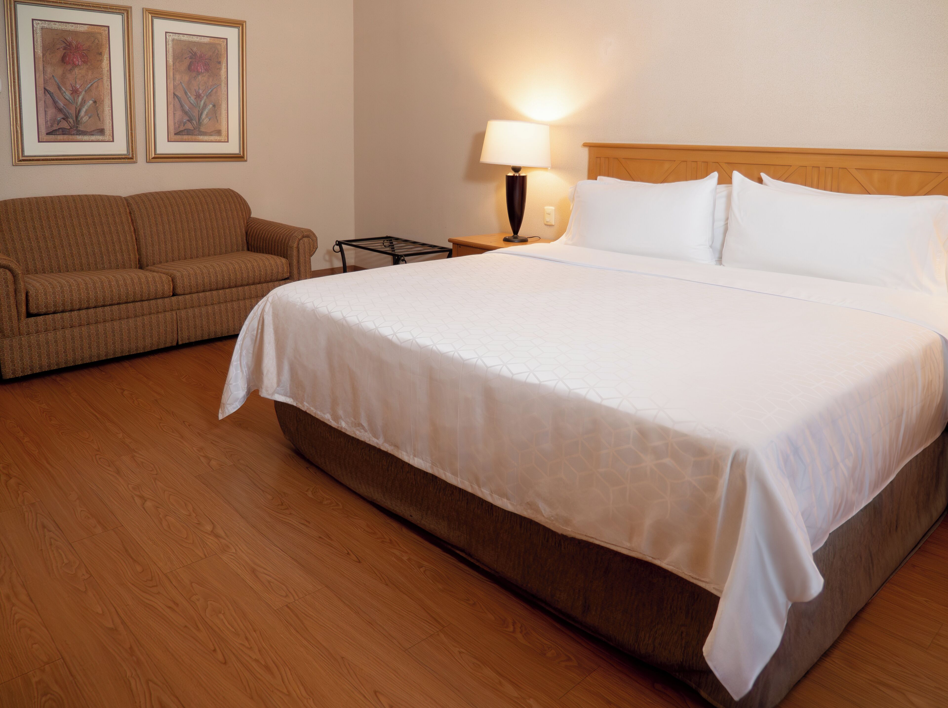 Habitación de hotel con una cama grande, un sofá, una mesita de noche con lámpara y dos cuadros en la pared.