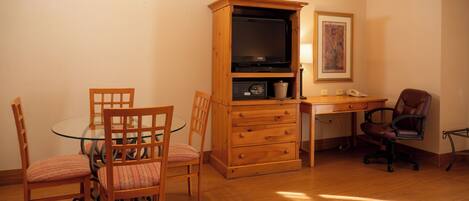 A dining area with a glass table and wooden chairs, a wooden cabinet with a television, and a desk with a chair.